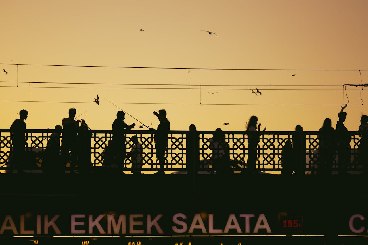 Silhouetted people on Galata Bridge in Istanbul during a vibrant sunset with birds flying overhead.