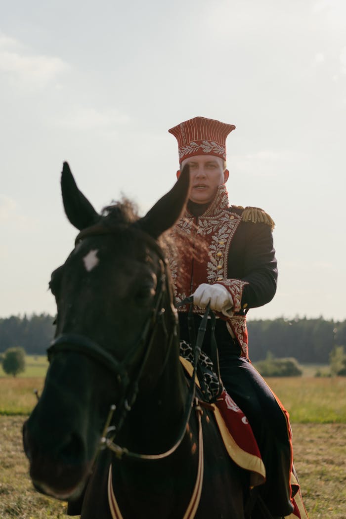 Man in historical military uniform on horseback outdoors against a pastoral backdrop.